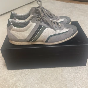 Grå och vita sneakers från Hugo Boss - Snygga grå och vita sneakers från Hugo Boss med snörning. Skorna har en stilren design med mörkare grå detaljer och en vit sula. Perfekta för en avslappnad och trendig look. Dom är i bra skick men smutsiga.