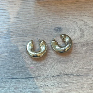 Guldiga örhängen i rund form - Snygga guldiga örhängen i en rund och tjock design. Perfekta för att ge en elegant touch till din outfit. De har en slät och blank yta som reflekterar ljuset fint.