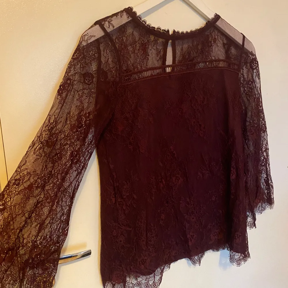 Säljer en elegant vinröd spetsblus med långa ärmar och vackra spetsdetaljer. Blusen har en rund hals och en liten knapp i nacken för stängning. Perfekt för en stilfull look.. Puserot.