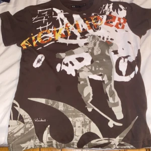 Brun t-shirt med skateboardtryck från F.L.Y. - (Stor i storlek, sitter som s) Cool brun t-shirt från F.L.Y. med ett stort skateboardtryck i vitt, grått och orange. Perfekt för den som gillar streetwear och skatekultur. T-shirten har korta ärmar och en avslappnad passform.