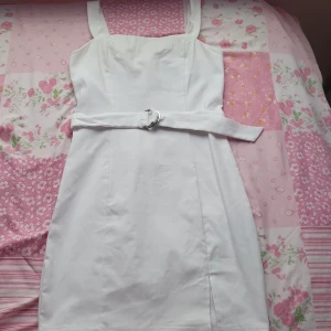 Vit klänning med bälte - Snygg vit klänning med breda axelband och ett matchande bälte i midjan. Klänningen har en stilren design med en liten slits framtill. Perfekt för somriga dagar eller speciella tillfällen, som till exempel studenten🇸🇪.