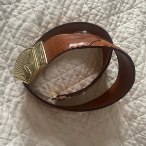 Brunt skärp med guldfärgat spänne - Snyggt brunt skärp i skinn med ett unikt guldfärgat spänne. Perfekt för att ge en stilfull touch till din outfit. Skärpet har en längd på 75-100 cm.