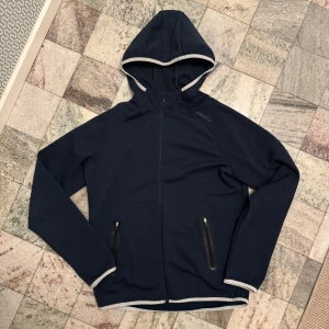 Mörkblå huvjacka från Craft - Säljer en mörkblå huvjacka från Craft i storlek M. Jackan har en dragkedja framtill och två fickor med dragkedjor. Den är långärmad och har ljusare kantdetaljer vid ärmslut och nederkant. Perfekt för en aktiv livsstil!