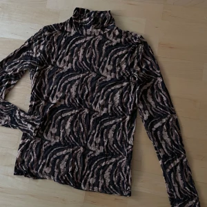 Mönstrad polotröja i brun och svart L-M stretch - Snygg polotröja med ett unikt mönster i beige och svart. Tröjan har långa ärmar och en hög krage, perfekt för att hålla värmen under kyligare dagar. Den är tillverkad i ett mjukt och bekvämt material som ger en skön passform.