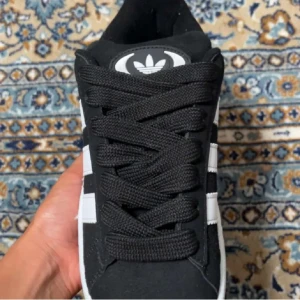 Svarta Adidas Campus sneakers - Säljer ett par svarta Adidas Campus sneakers i nyskick. Skorna har den klassiska tre-randiga designen i vitt och är gjorda i skinn. De kommer med extra vita skosnören. Perfekta för både vardag och fest!