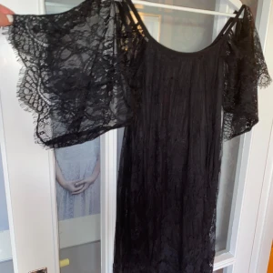 Svart spetsklänning - Säljer en elegant svart klänning med vackra spetsdetaljer. Klänningen har korta ärmar och en lös passform, perfekt för festliga tillfällen. Materialet är lätt och luftigt, vilket gör den bekväm att bära.