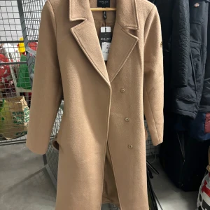 Beige kappa från Hollies - Säljer en superfin beige kappa från Hollies i storlek 36. Den är långärmad och har en klassisk design med knappar framtill. Perfekt för höst och vår, och den har ett bälte i midjan för en snygg passform. Materialet känns lyxigt och håller dig varm under kyliga dagar.