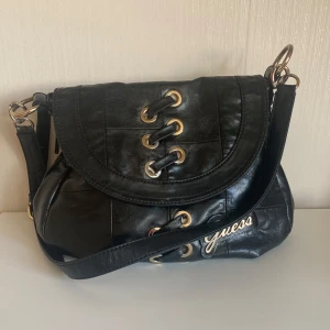 Svart axelväska från Guess - Säljer en snygg svart axelväska från Guess i skinn. Väskan har coola snördetaljer på framsidan och en praktisk dragkedja på baksidan. Insidan har tyvärr fläckar från kulspetspenna men det är inget man tänker på. Perfekt för både vardag och fest! 