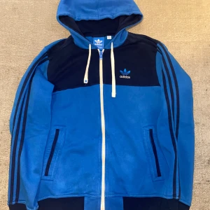 Blå och svart hoodie från Adidas - Säljer en snygg blå och svart hoodie från Adidas. Den har en dragkedja framtill och klassiska Adidas-ränder längs ärmarna. Perfekt för både träning och vardag. Huvan har snören för justering och det finns två praktiska fickor framtill.