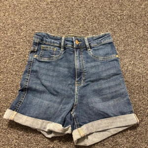 Blå jeansshorts från Gina Tricot - Snygga blå jeansshorts från Gina Tricot med hög midja och uppvikta ben. Perfekta för en avslappnad sommarlook. Klassisk femficksdesign och knappgylf.