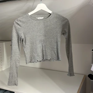 Grå ribbad tröja från MANGO - Säljer en stilren grå ribbad tröja från MANGO. Tröjan har långa ärmar och en croppad design med vågiga kanter vid ärmslut och nederkant. 