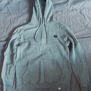 Grön hoodie från Les Deux - Säljer en stilren gröngrå hoodie från Les Deux med justerbar huva och känguruficka. Perfekt för en avslappnad look. Den har en liten logga på bröstet och ribbade muddar vid ärmslut och nederkant. Den har två väldigt små hål på ryggen, men absolut inget som märks.