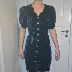 Mörkgrön sammet klänning med knappar - Elegant vintage mörkblå klänning i sammet med korta puffärmar och dekorativa knappar framtill. Perfekt för en stilfull look. Köpt second hand, aldrig använd av mig. Helt i nyskick förutom lite slitage på knapparna längst ner men det är inget som syns när den är på. Det gör ont i mig att sälja den här!! Men den passar tyvärr inte.