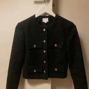 Svart cardigan med knappar i boucle  - Elegant svart kavaj med dekorativa knappar framtill. Den har en klassisk design med rund hals och långa ärmar. Perfekt för en stilren look.