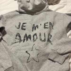 Grå hoodie med tryck - Säljer en grå hoodie med texten 'Je m'en amour' och en stjärna tryckt på framsidan. Från gina