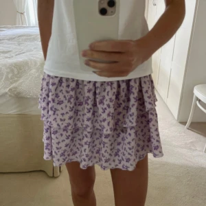 Lila blommig kjol - Söt lila kortkjol med blommigt mönster och volangdetaljer. Perfekt för en somrig look. Den elastiska midjan ger en bekväm passform. M men passar S