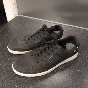 Svarta sneakers från Jack & Jones - Snygga svarta sneakers från Jack & Jones med vit sula och snörning. Skorna har en stilren design med diskreta detaljer. Använt fåtal gånger. Tvättar såklart skorna innan dem skickas iväg.