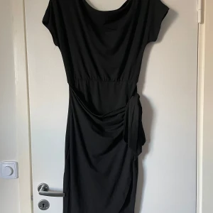 Svart klänning med knyt - Elegant svart klänning med knyt i midjan. Den har en smickrande passform och korta ärmar. Perfekt för en stilren look.