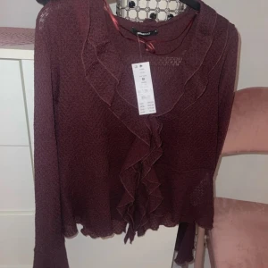 Vinröd blus med volangdetaljer - Säljer en elegant vinröd blus från gina med volangdetaljer framtill. Blusen har långa ärmar och en fin struktur i tyget. Perfekt för att ge en stilfull touch till din outfit.