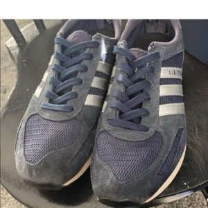  Adidas La trainers - Snygga blå sneakers från Adidas med klassiska ränder i silver. Skorna har en sportig design med snörning och en bekväm sula. Perfekta för en casual look.