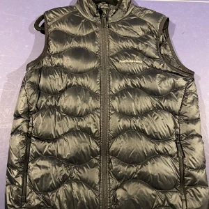 Svart väst från Peak Performance - Snygg svart dunväst från Peak Performance i storlek 170 med dragkedja framtill. Västen har en quiltad design som ger extra värme och stil. Perfekt året runt och passar till allt!