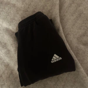 Svarta träningsbyxor från Adidas - Snygga svarta träningsbyxor från Adidas med klassiska vita ränder längs sidorna. Byxorna har en elastisk midja för extra komfort och är perfekta för sport och träning.