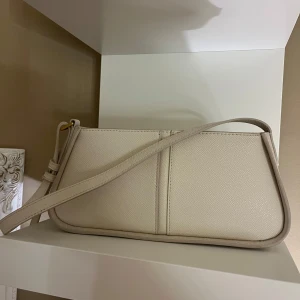 Beige axelväska - Snygg beige axelväska i syntetmaterial med enkel och stilren design. Väskan har en justerbar axelrem och en praktisk storlek för vardagsbruk. 