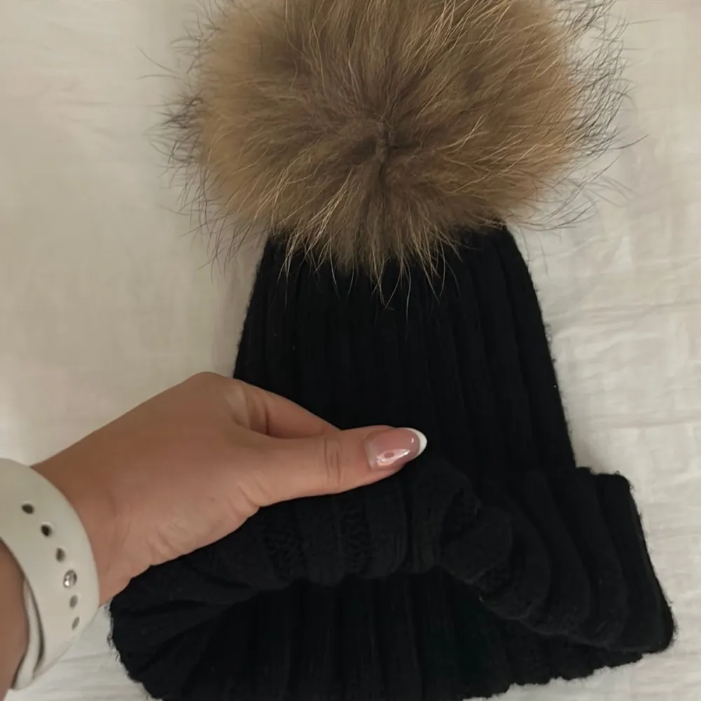 Snygg svart ribbad mössa med en stor beige pälsboll på toppen. Perfekt för att hålla värmen under kalla dagar. Stilren och enkel design som passar till det mesta.. Asusteet.