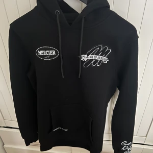Svart hoodie från Mercier UK - Snygg svart hoodie från Mercier UK med tryck på baksidan.  Den har en klassisk känguruficka och justerbar huva med dragsko. Perfekt för en avslappnad stil.