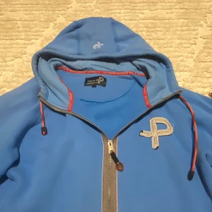 Blå huvtröja från Peak Performance - Snygg blå huvtröja från Peak Performance med dragkedja och broderad logga på bröstet. Tröjan har långa ärmar och justerbar huva med dragsko. Perfekt för en avslappnad stil.