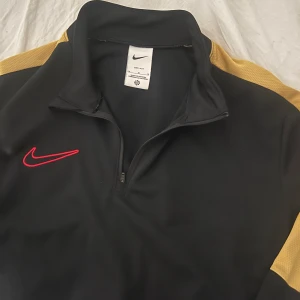 Svart och gul tröja från Nike - Snygg svart tröja från Nike med gula detaljer på ärmarna och en röd Nike-logga på bröstet. Tröjan har en dragkedja vid halsen och långa ärmar, perfekt för sportiga aktiviteter.