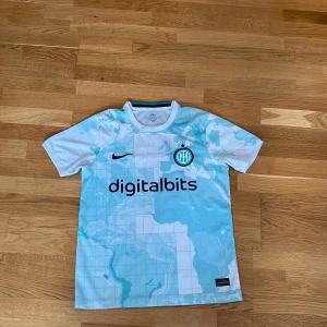 Inter fotbollströja - Säljer en snygg vit och ljusblå fotbollströja från Nike med ett världskartemönster. Tröjan har korta ärmar och tryck från Digitalbits och Lenovo. Perfekt för fotbollsfans! Den är extremt skön och passar folk mellan 174-183.