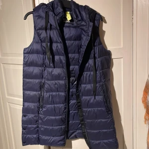 Mörkblå dunväst från C&A - Snygg och praktisk mörkblå dunväst från C&A. Västen har en huva och dragkedja framtill, perfekt för kyliga dagar. Den quiltade designen ger en stilren look.