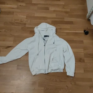 Vit hoodie med dragkedja - Säljer en stilren vit hoodie med dragkedja och huva. Tröjan har långa ärmar och ribbade muddar vid ärmslut och nederkant. Perfekt för en avslappnad look. Tröjan har en liten fläck vid armen. Har använt 2-3 gånger. Obs ej original 