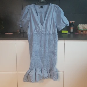 Ljusblå klänning från Gina Tricot - Charmig ljusblå klänning från Gina Tricot med v-ringning och korta puffärmar. Klänningen har en smockad midja och volangdetalj vid fållen, vilket ger en lekfull touch. Perfekt för vår och sommar!