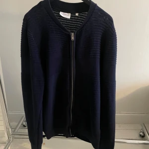 Mörkblå kofta från Lindbergh - Snygg mörkblå kofta från Lindbergh med dragkedja framtill. Den har en ribbad struktur och långa ärmar, perfekt för kyligare dagar. Enkel och stilren design som passar till många tillfällen.