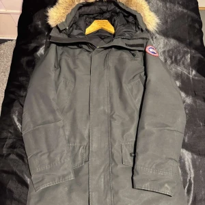 Canada Goose jacka (ÄKTA) - Säljer en stilren grå parkajacka från Canada Goose med pälsdetaljer på huvan. Jackan har en broderad logotyp på ärmen och praktiska fickor framtill. Perfekt för kalla dagar.