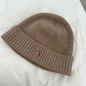 Beige stickad mössa från Tommy Hilfiger - Snygg beige stickad mössa från Tommy Hilfiger med ribbad kant. Perfekt för kyliga dagar med stilren design och diskret logotypdetalj framtill.
