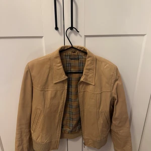 Beige skinnjacka  - Snygg beige skinnjacka med klassiskt rutigt foder. Jackan har en dragkedja framtill och ribbade muddar vid ärmslut och nederkant. Perfekt för en stilren look.Liknar Burberry för dem som inte vill slösa massor med pengar på burberry är den här perfekt 👌 