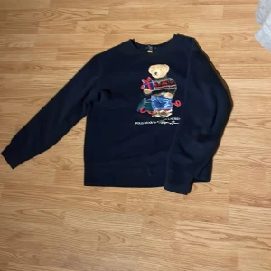 Polo Ralph Lauren tröja (14-16) - Säljer en mörkblå tröja från Polo Ralph Lauren med ett gulligt nallemotiv på framsidan. Tröjan har långa ärmar och en klassisk rund halsringning. Perfekt för en avslappnad stil med en touch av lekfullhet. Storlek 158 -164  skick 7/10