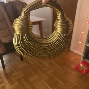Guldig handväska i unik design - Säljer en unik guldig handväska med en iögonfallande design. Väskan har en rundad form med flera lager av guldfärgat material som ger en elegant och modern look. Perfekt för att sticka ut på festen eller andra speciella tillfällen.