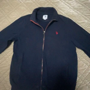 Mörkblå tröja med dragkedja från U.S. Polo Assn. - Säljer en stilren mörkblå tröja från U.S. Polo Assn. med dragkedja och röd logga på bröstet. Perfekt för en avslappnad look. Tröjan har långa ärmar och en bekväm passform.