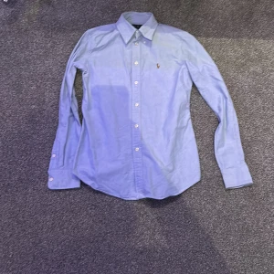 Blå skjorta från Ralph Lauren - Snygg blå skjorta från Ralph Lauren med klassisk krage och broderad logga på bröstet. Skjortan har långa ärmar och knappar framtill. Perfekt för en stilren look.