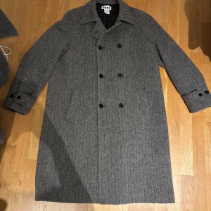 Grå kappa från Hope - Snygg grå kappa från Hope i klassiskt fiskbensmönster. Dubbelknäppt design med långa ärmar och stiliga knappar. Perfekt för kyligare dagar med sin eleganta och tidlösa stil.