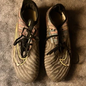 Nike phantom fotbollsskor  - Snygga fotbollsskor från Nike med ett unikt mönster i flera färger. Skorna har snörning och en slitstark sula med dobbar, perfekt för spel på gräs. De har en modern design med en mix av färger som ger ett coolt intryck. 