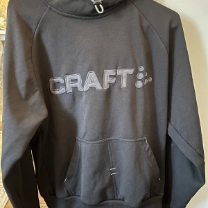 Svart hoodie från Craft - Snygg svart hoodie från Craft med stor logga på bröstet. Den har en praktisk känguruficka och en justerbar huva med dragsko. Perfekt för en avslappnad stil.