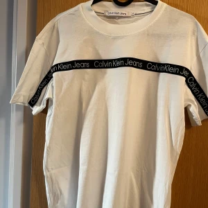 Vit t-shirt från Calvin Klein Jeans - Snygg vit t-shirt från Calvin Klein Jeans med svart logotypband över bröstet. Perfekt för en stilren och modern look. Kortärmad och tillverkad i mjukt material för extra komfort.