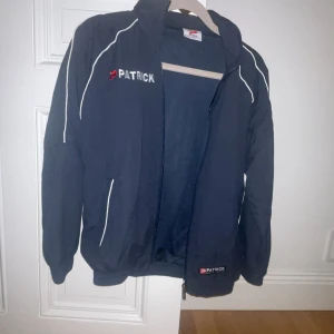 Svart vindjacka från Patrick - Snygg svart vindjacka från Patrick med vita detaljer och dragkedja. Perfekt för blåsiga dagar. Jackan har en sportig look med broderad logga på bröstet. SÅ snygg men säljer då den är lite för liten på mig som är strl S.