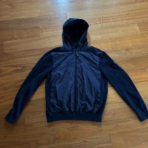 Mörkblå cardigan från Canada Goose - Snygg mörkblå cardigan från Canada Goose med huva och dragkedja. Jackan har ett stilrent märke på ärmen och är perfekt för kyligare dagar. Den är köpt från instasida och använd Max 10 gånger. Skick 10/10. Storlek L men passar mer som M/S