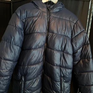 Blå pufferjacka från 11Project - Säljer en blå pufferjacka från 11Project. Jackan har en dragkedja framtill och en huva för extra skydd mot väder. Perfekt för kyligare dagar❄️Skriv vid minsta fundering!😁